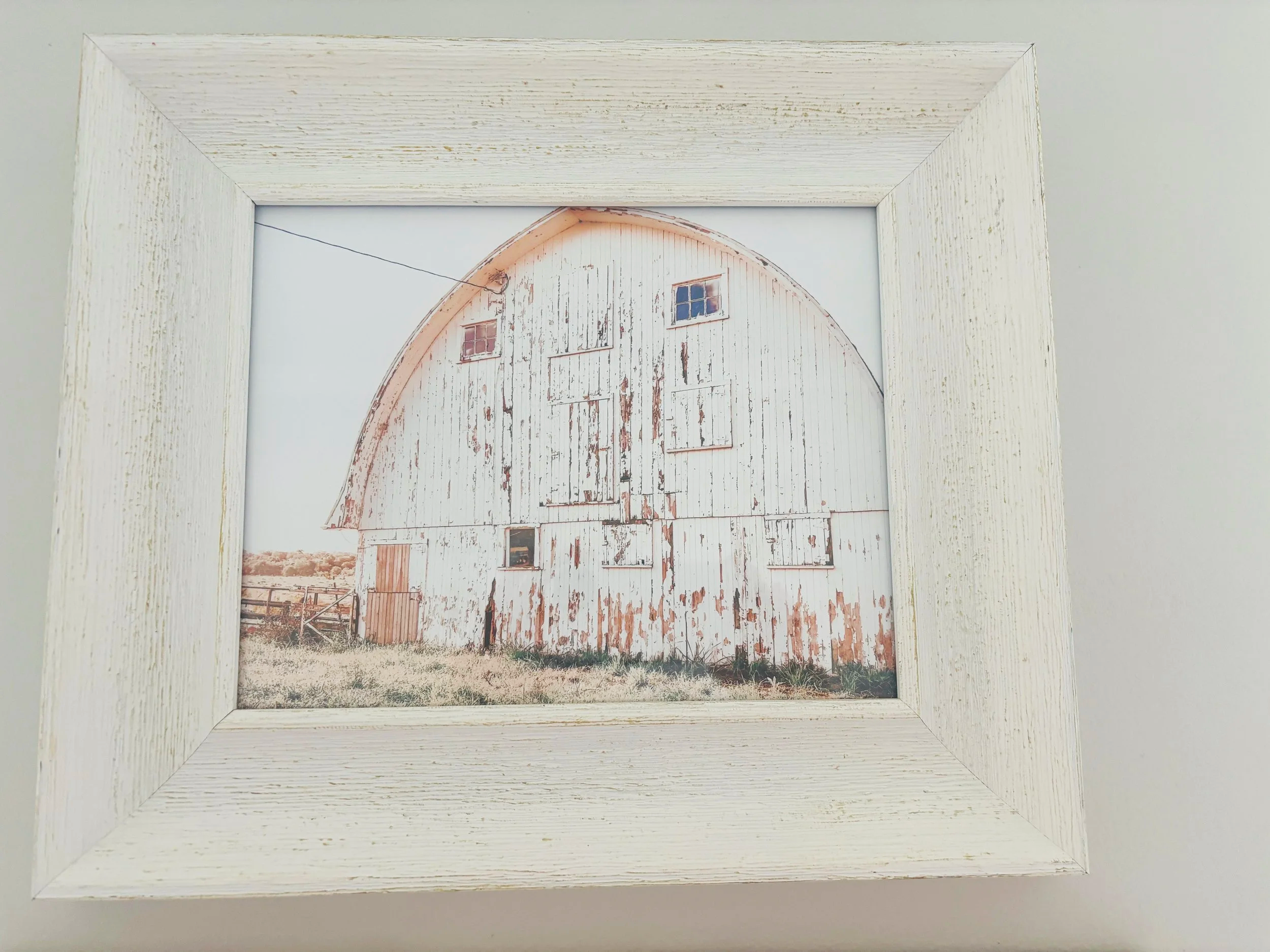 A framed picture of an old, weathered barn with peeling white paint, two small windows, and a large sliding door, set in a rural landscape with a grassy field and wooden fence.