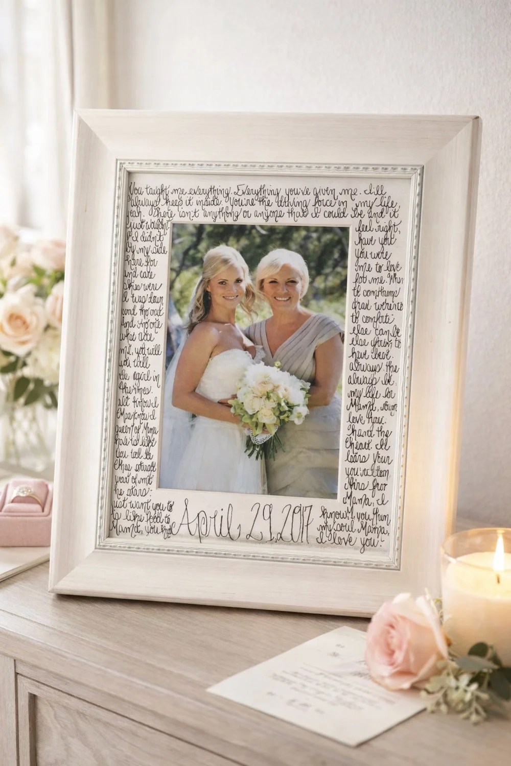 A framed wedding photo of two women, one in a wedding dress, surrounded by handwritten notes and the date April 29, 2017, on a light wooden table along with a lit candle, pink roses, and a small pink box.