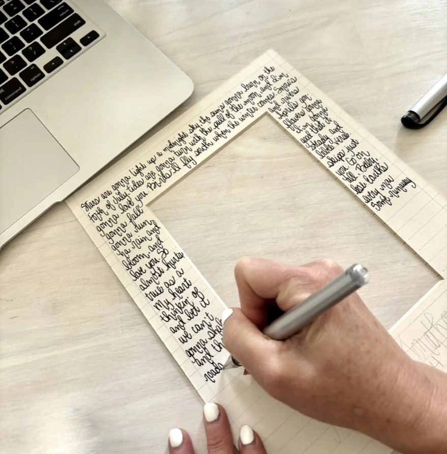 A person is writing in a notebook with lined pages, surrounded by a silver marker and a silver laptop on a wooden surface.