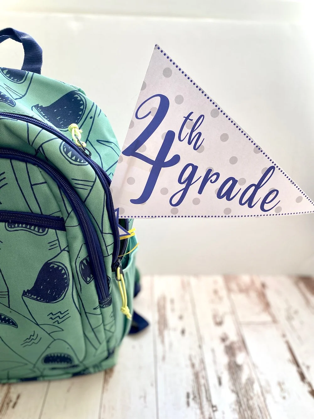 A green backpack with dark blue zippers and a yellow zipper pull, next to a sign that reads '24th grade,' on a wooden floor.