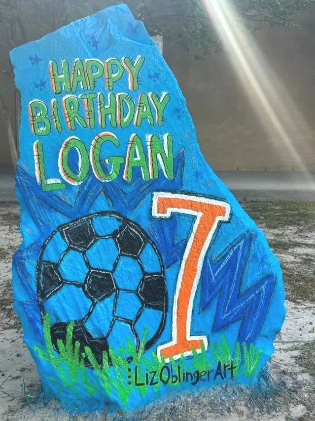 Colorful painted rock with a birthday message reading "Happy Birthday Logan," a soccer ball, the number 7, and the signature LizOblingerArt in green grass.