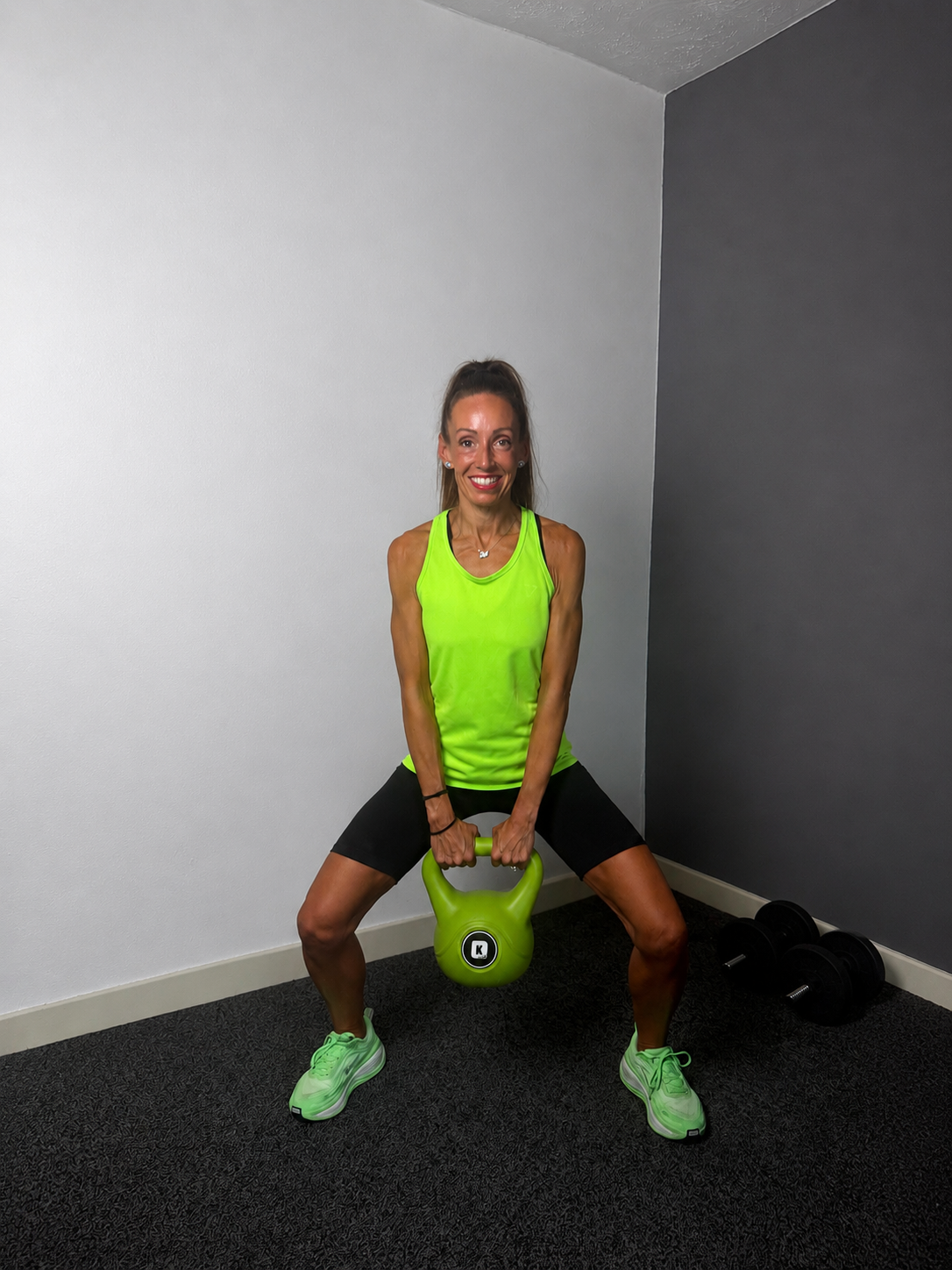 Woman in neon green tank top and black shorts holding a kettlebell in a fitness room, smiling at the camera.
