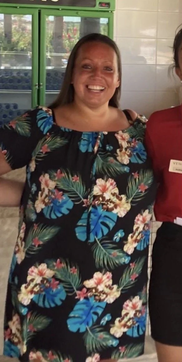 A woman with long brown hair smiling, wearing a black dress with a colorful floral and leaf pattern, standing indoors near a green glass display or refrigerator.