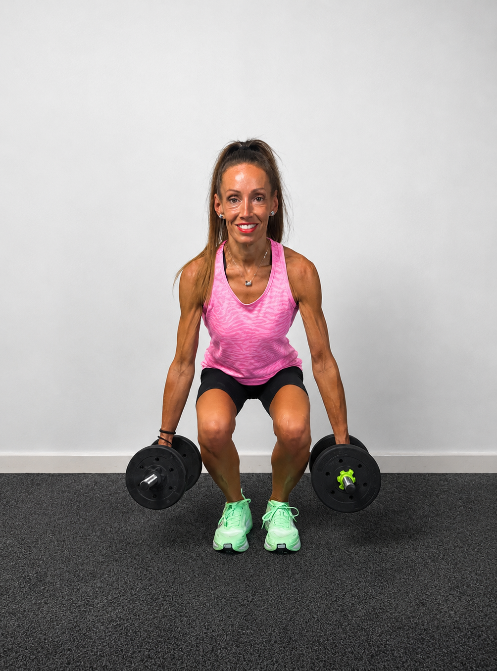 Woman exercising with dumbbells indoors, wearing pink tank top, black shorts, and green sneakers, against a plain white wall.