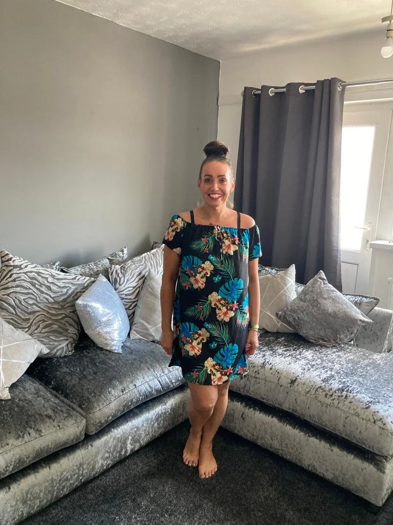Woman standing barefoot on a carpeted floor in a living room with a large sectional sofa, pillow cushions, gray curtains, and a window in the background.