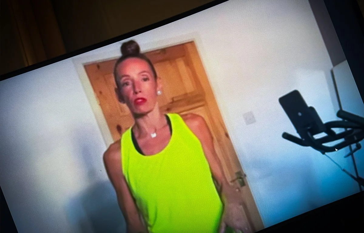 A woman with red hair tied in a bun, wearing a neon green tank top, standing indoors in front of a wooden door, with a treadmill partially visible on the right side of the image.