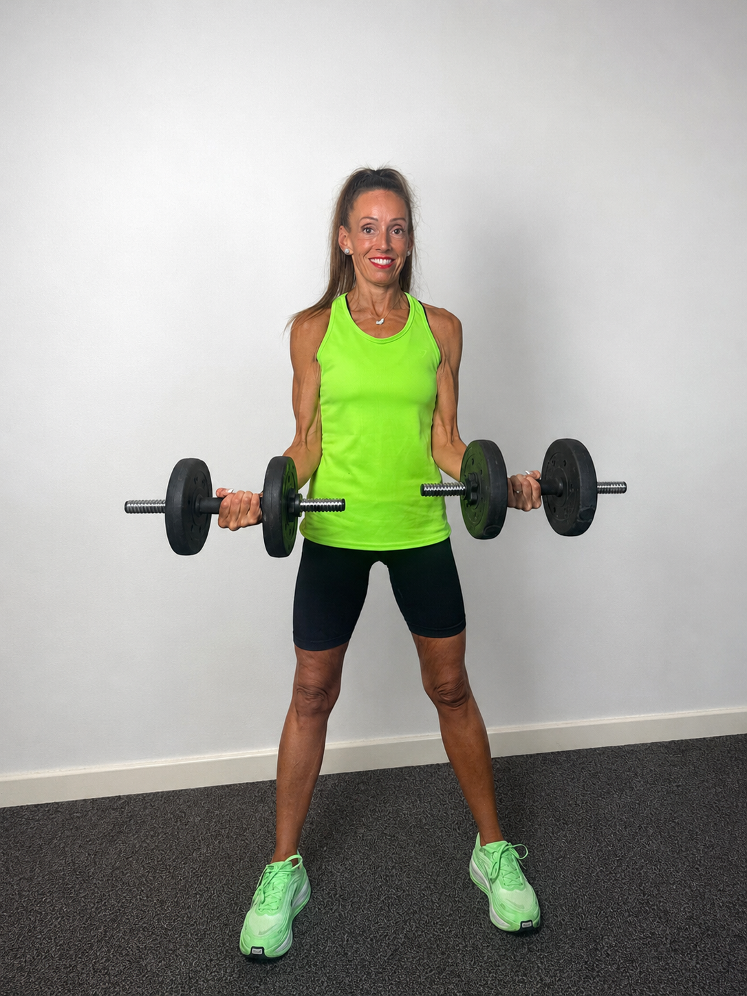 An older woman in a bright green athletic top and black shorts lifting dumbbells in a gym.