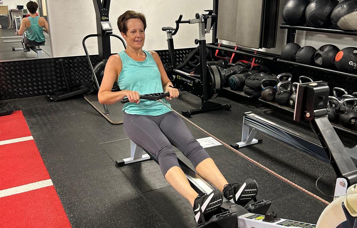 A woman using a rowing machine in a gym, with gym equipment on shelves behind her.