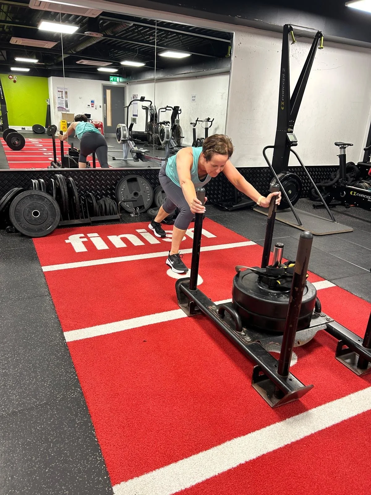 A woman exercising with a sled at the gym, pulling weights on a test rig. She is wearing a turquoise shirt and black shorts, with a mirror to her left reflecting her activity. The gym has various equipment and a red mat with white text.