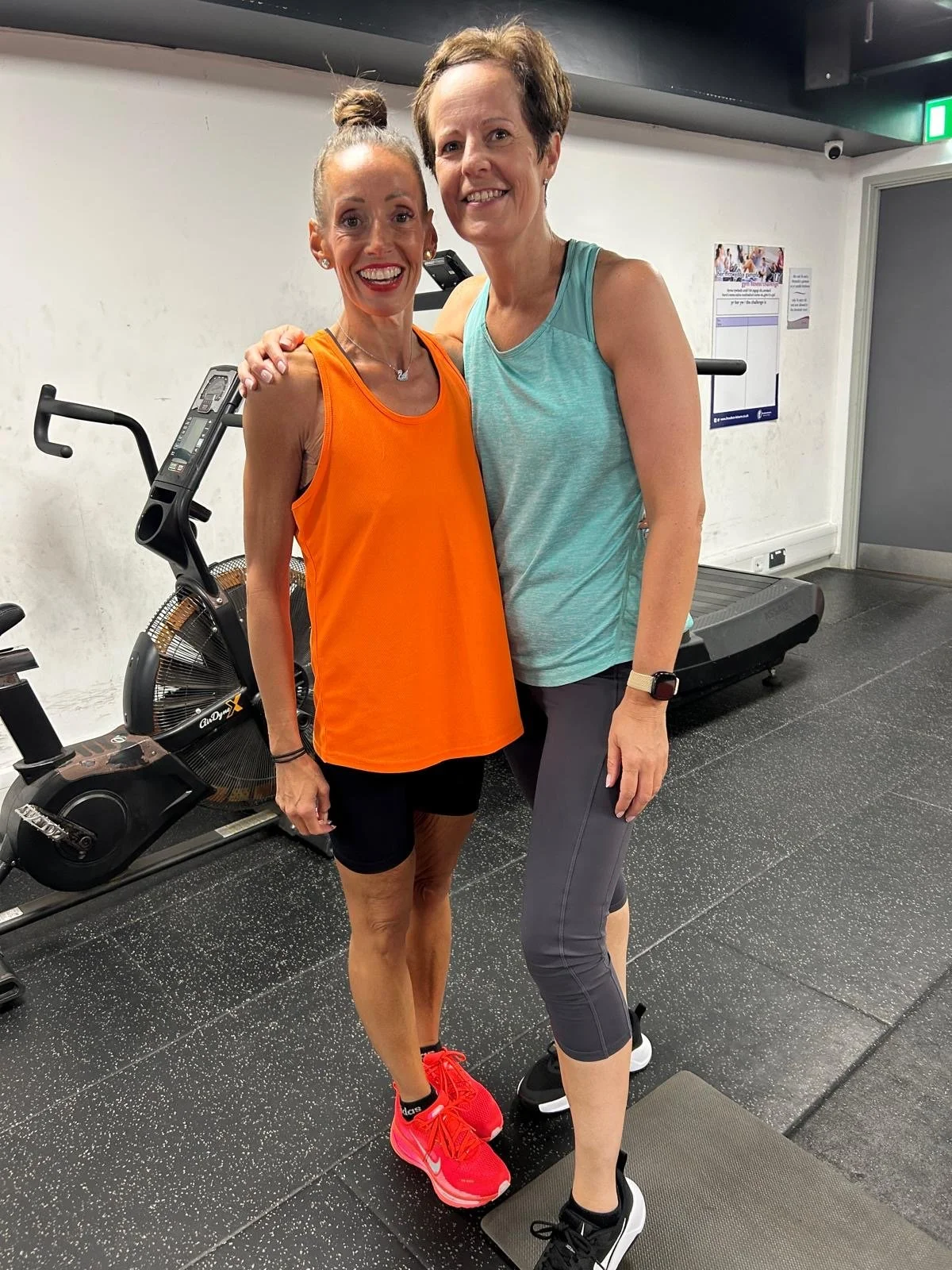 Two women standing together in a gym, smiling. One woman is wearing an orange sports top, black shorts, and bright pink running shoes. The other woman is wearing a light teal tank top, dark gray leggings, and black sneakers. There is exercise equipment, a treadmill, and a poster on the wall in the background.