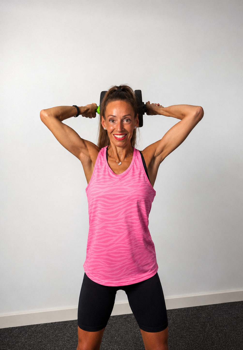 A smiling woman with long brown hair in a ponytail, wearing a pink athletic tank top and black shorts, holding a resistance band behind her neck with both hands, standing in an indoor setting.