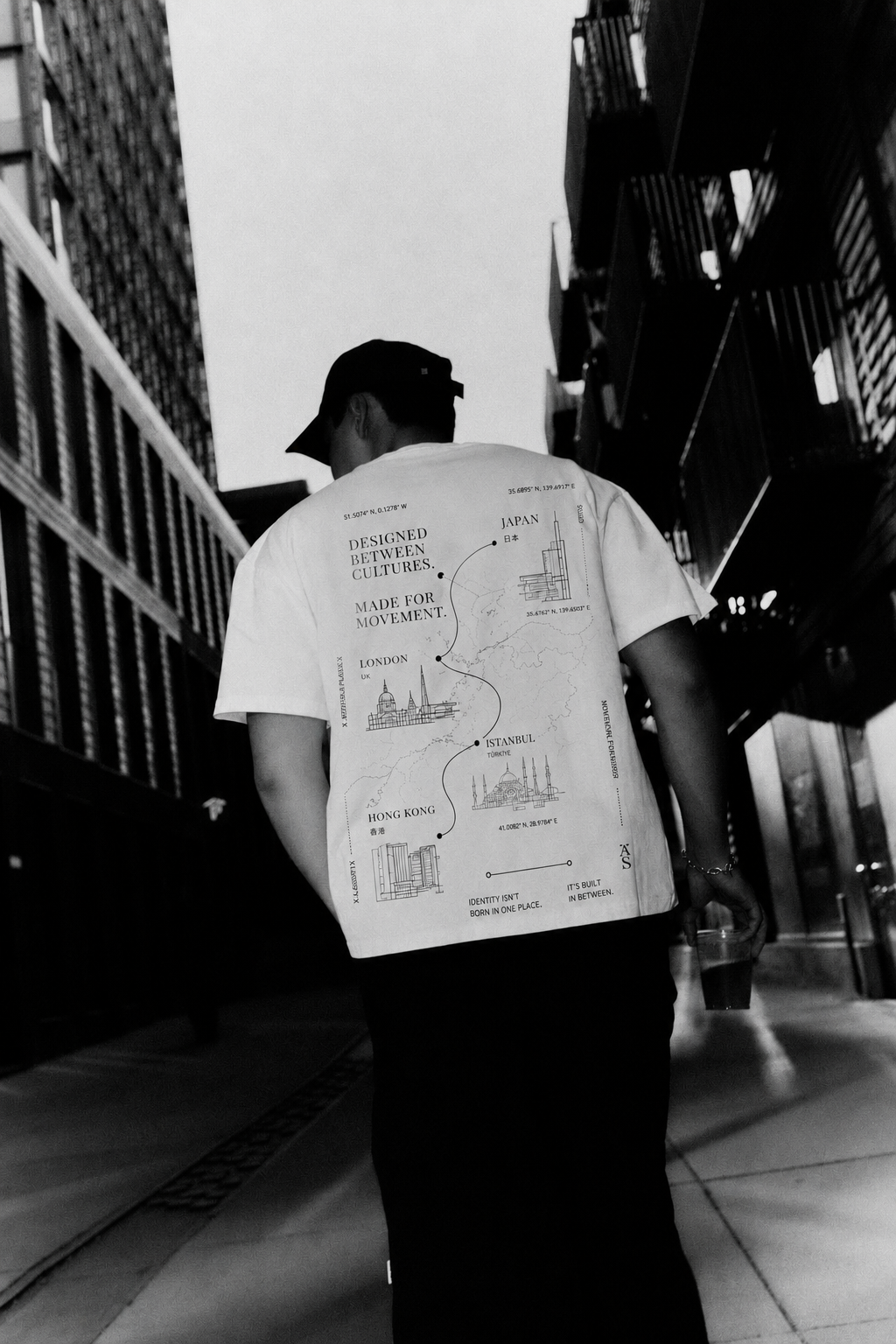 A person walking down a city street at night, wearing a t-shirt with a world map highlighting cities like London, Istanbul, Hong Kong, and Japan, with the text 'Designed between cultures. Made for movement.'