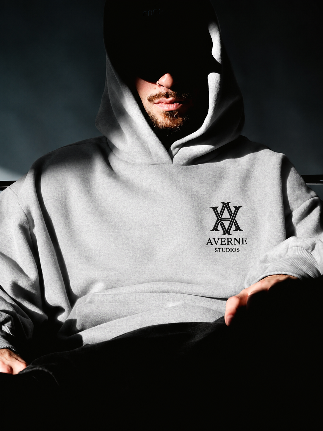 A man wearing a light gray hoodie with a logo reading 'AVENUE STUDIOS' on the front, sitting with his head slightly bowed and his face mostly obscured in shadow, creating a mysterious and dark atmosphere.