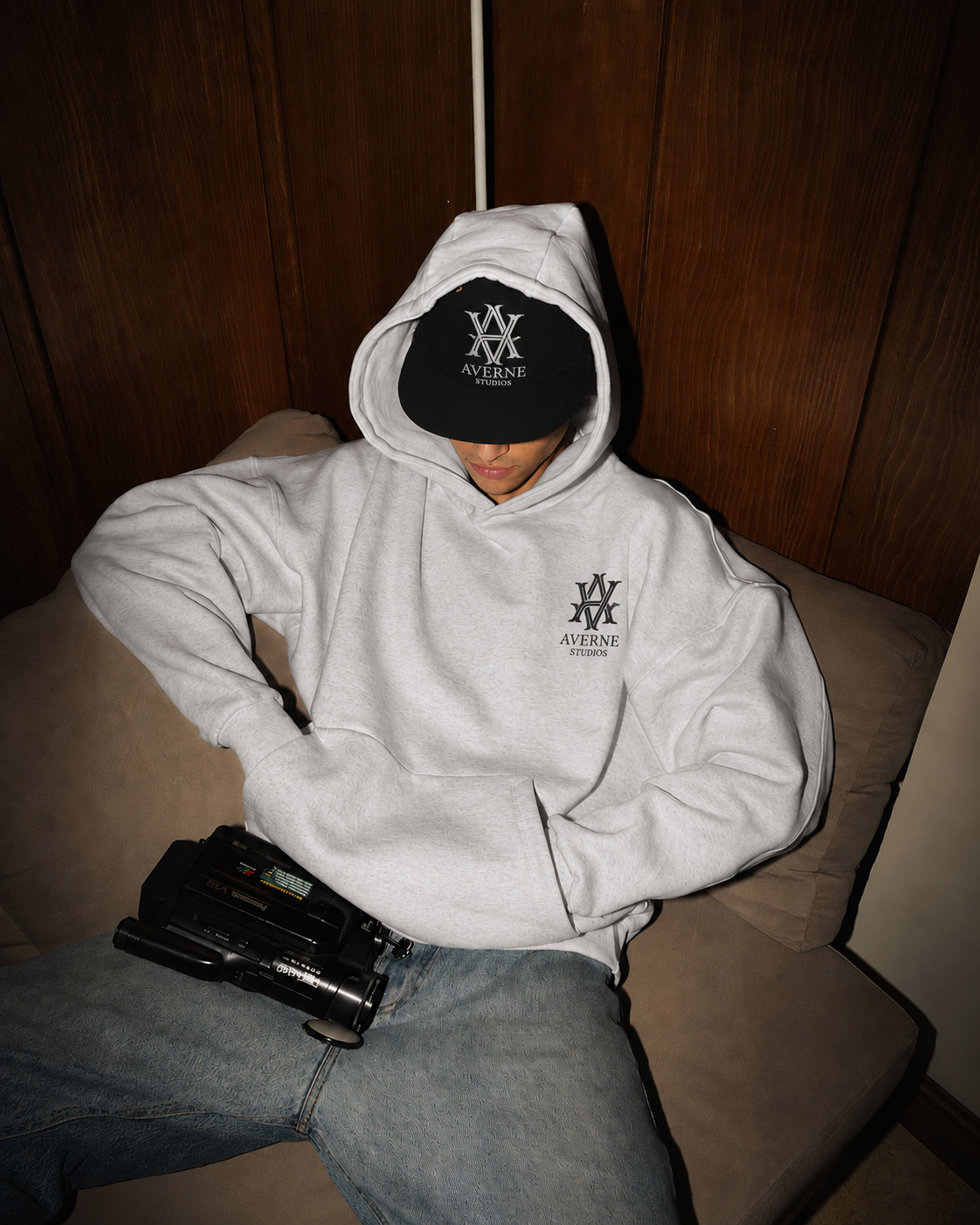 A young man sitting on a beige sofa, wearing a white hoodie with the logo 'Averne Studios,' a black cap with the same logo, and blue jeans. He has a vintage camcorder on his lap, with his head slightly bowed and face partially obscured by the cap. Th
