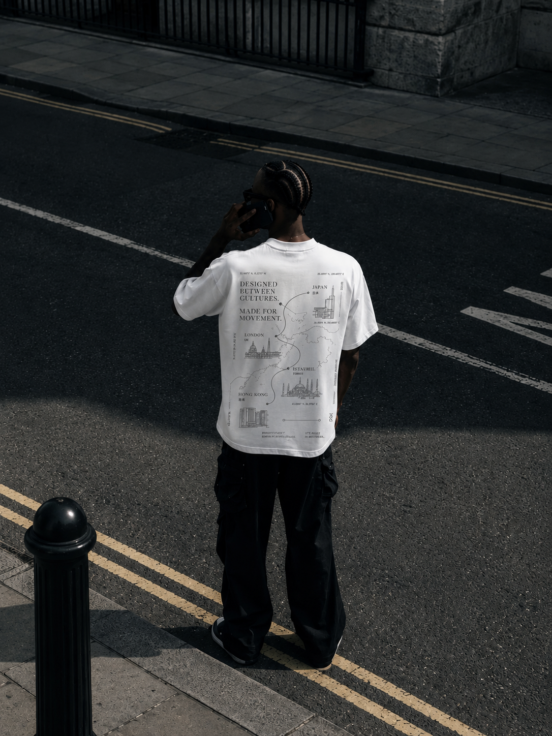 A person standing on a city street curb wearing a white oversized t-shirt with world maps and landmarks design, talking on a cell phone, with dark pants and cornrow hairstyle.