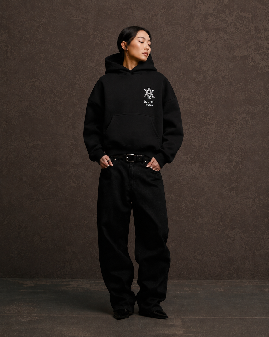 A woman in black hoodie and pants standing against a brown textured background, looking to her right with hands in her pockets.