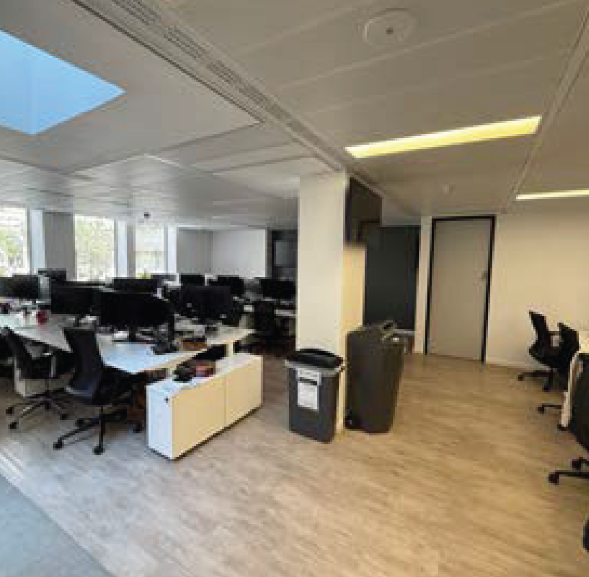 Modern office space with rows of desks and black office chairs, large windows letting in natural light, ceiling with air vents, and fluorescent lighting.