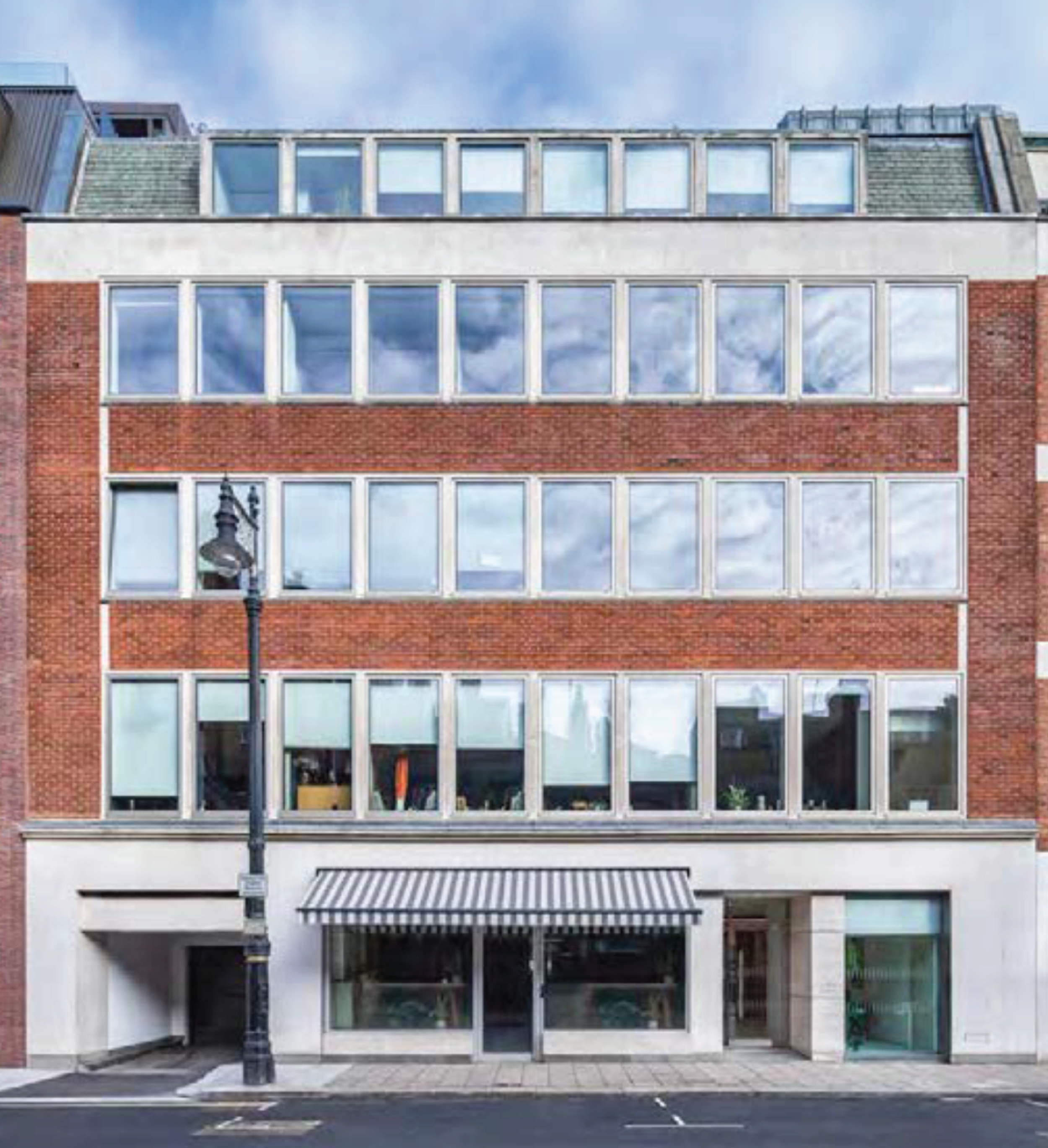 Multi-story modern building with large glass windows and a red brick facade. There is a street lamp and a striped awning in front of the building, with a cloudy sky reflected in the windows.