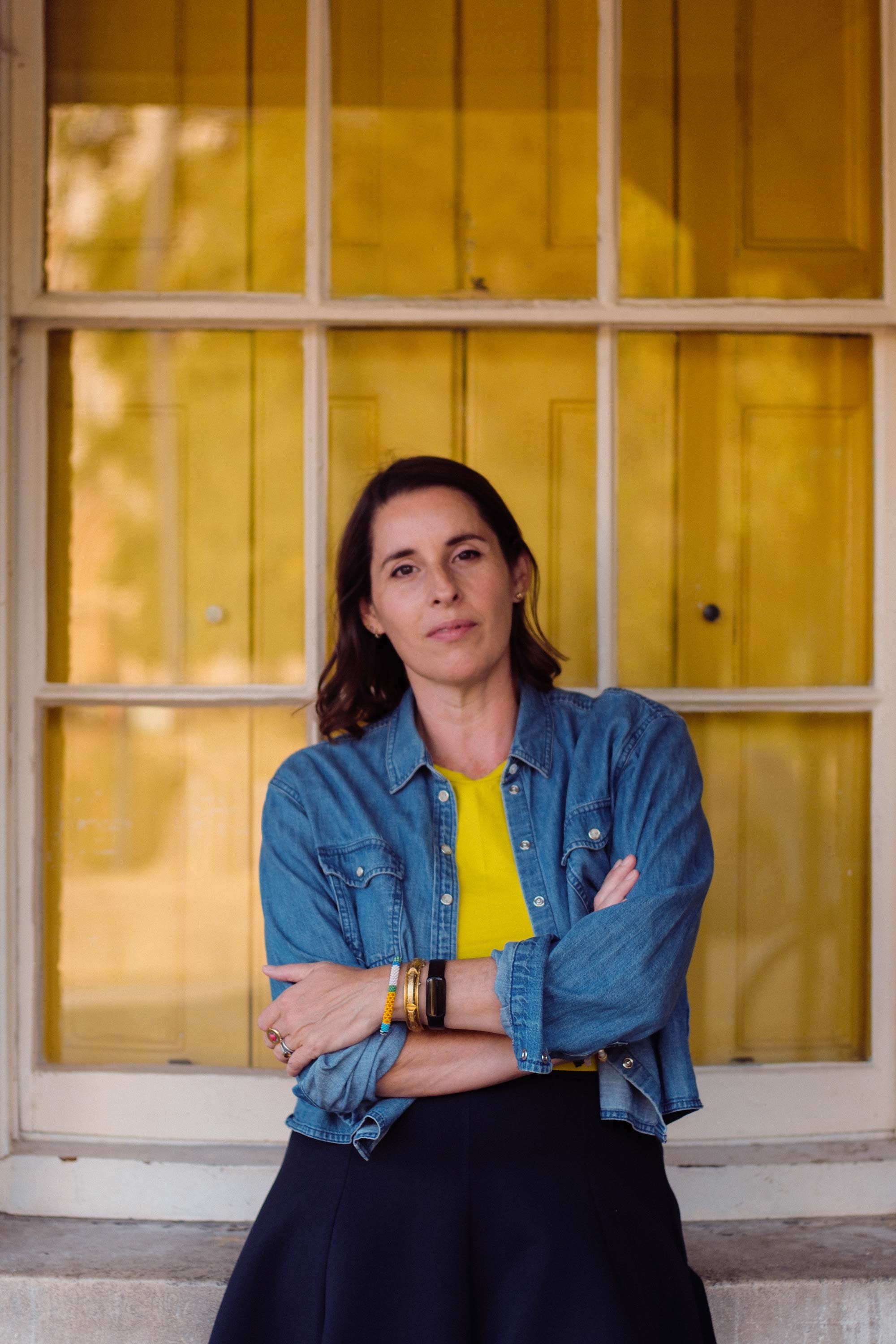 A woman with dark brown hair wearing a denim jacket and a yellow shirt posing with arms crossed in front of a yellow door or window frame.