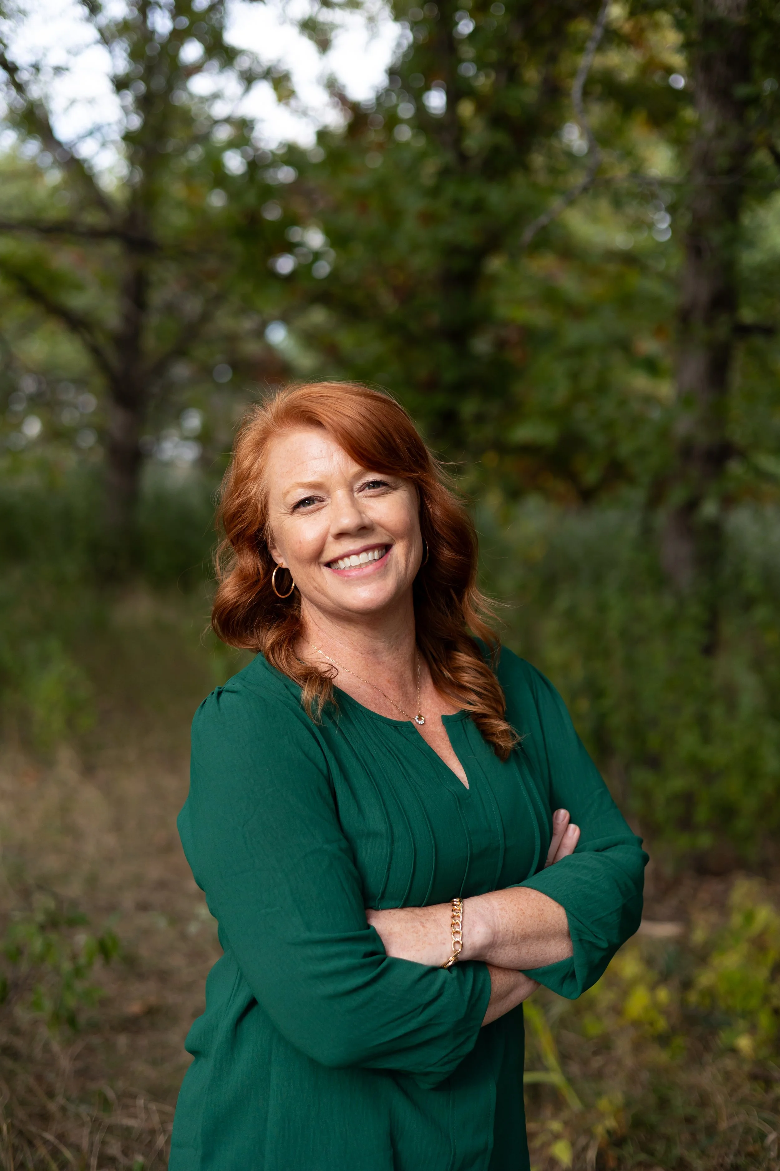 A woman with red hair smiling outdoors with her arms crossed, wearing a green top and jewelry, in front of a background of trees and greenery.