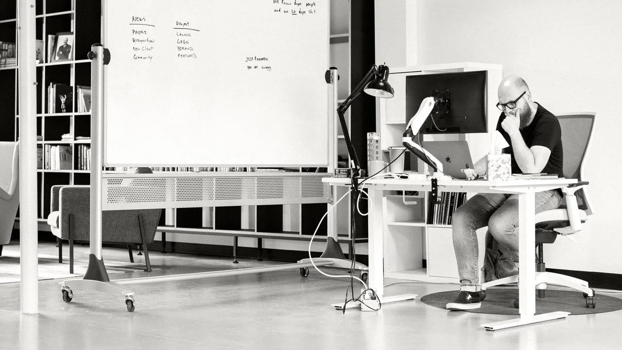 Jon Eslinger, a man with glasses and a beard working at a desk with a laptop, tissue box, and desk lamp in an office. There is a whiteboard with writing and a bookshelf with framed photos and books in the background.