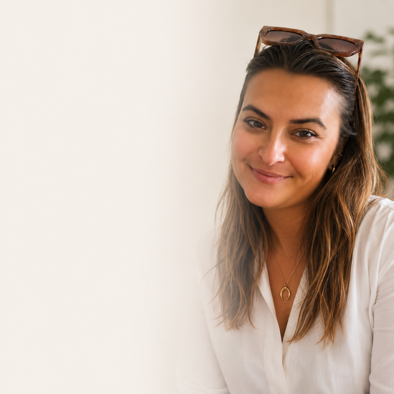 A smiling woman with brown hair wearing sunglasses on her head, a white shirt, and gold jewelry, sitting indoors with a light-colored background and a blurred plant in the distance.