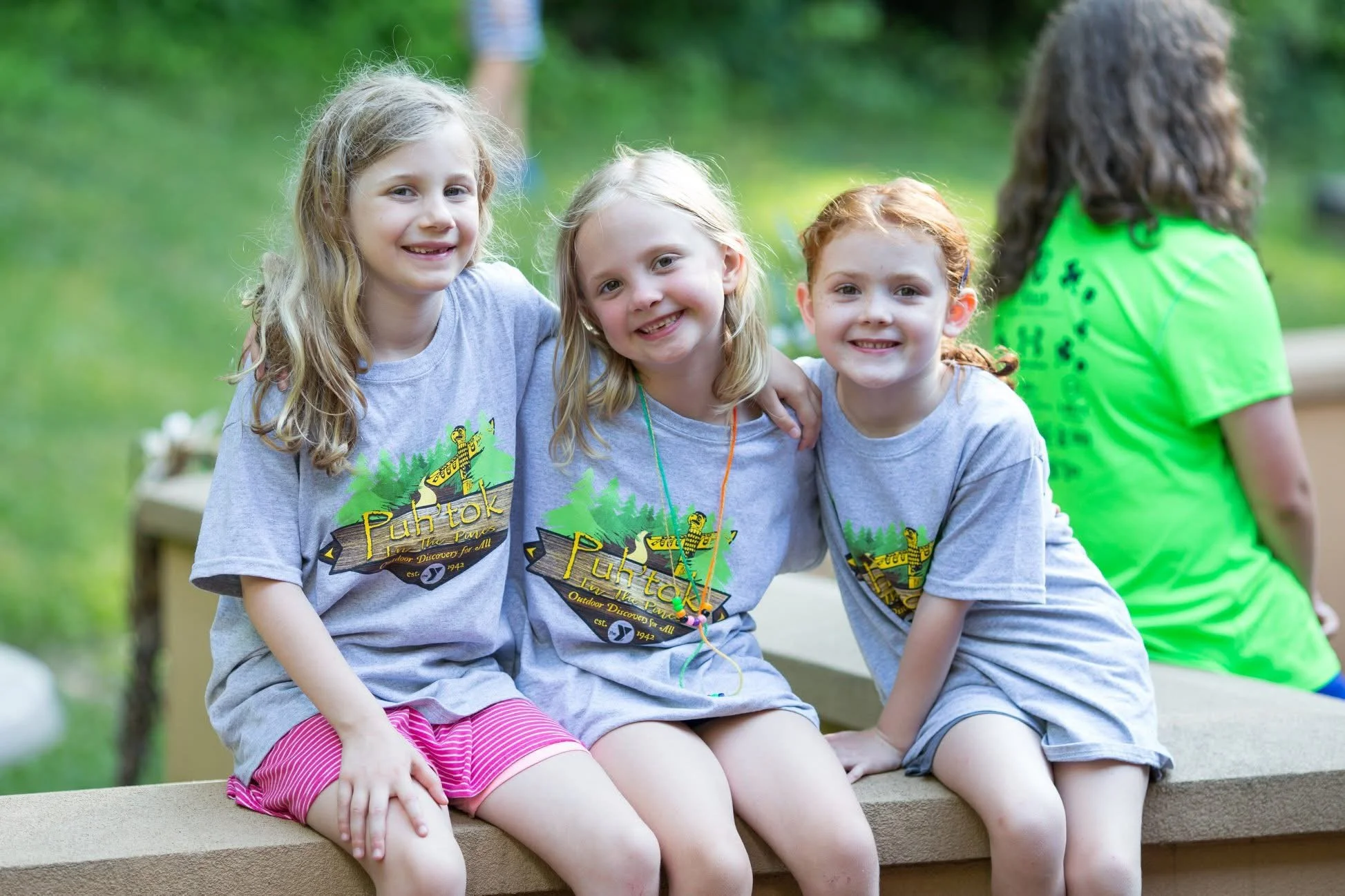 Three young girls sitting on a concrete ledge outdoors, smiling at the camera, wearing gray t-shirts with a yellow and green camp logo, with two more children partially visible in the background.