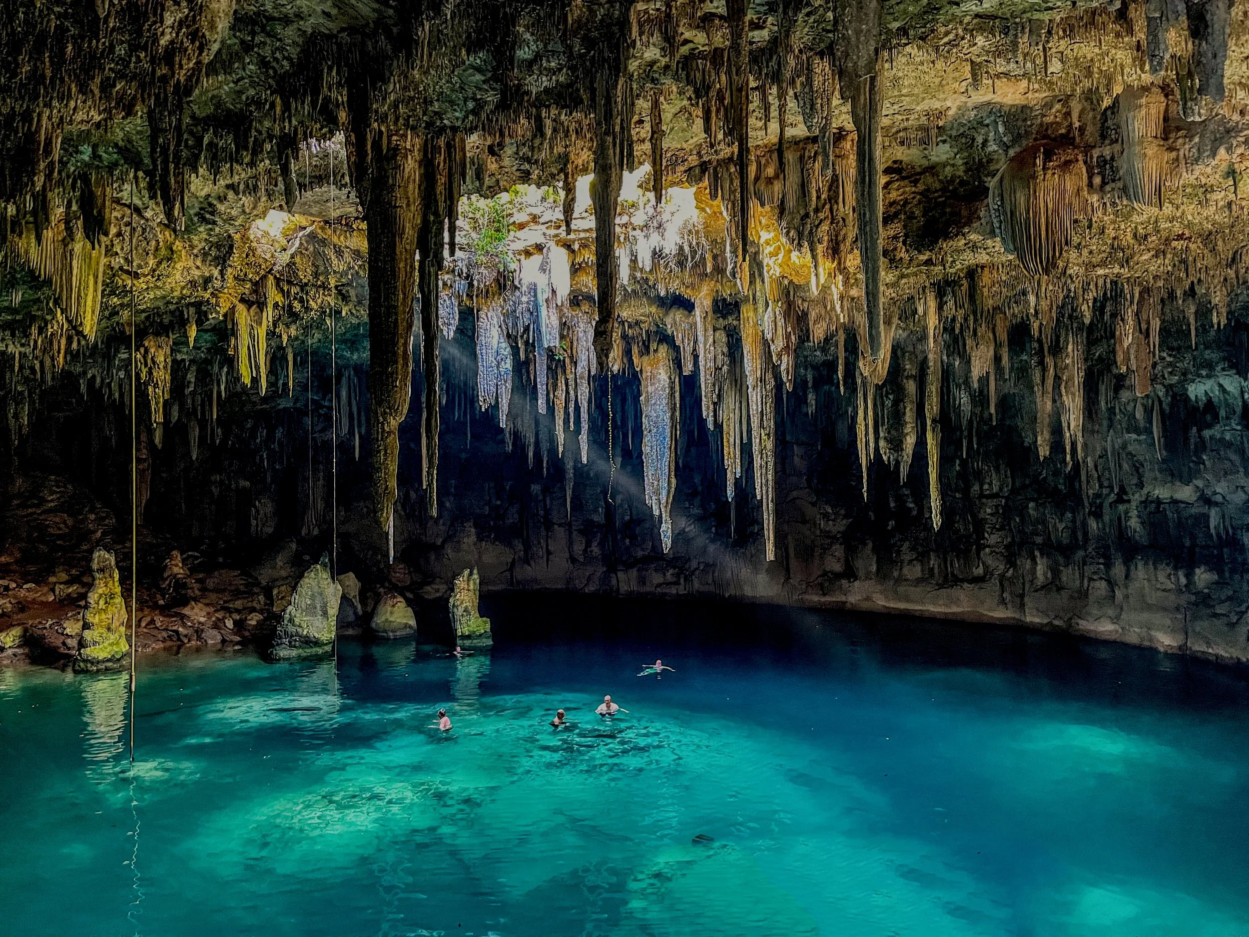 Swimmers enjoy a turquoise lake inside a large cave with stalactites hanging from the ceiling.