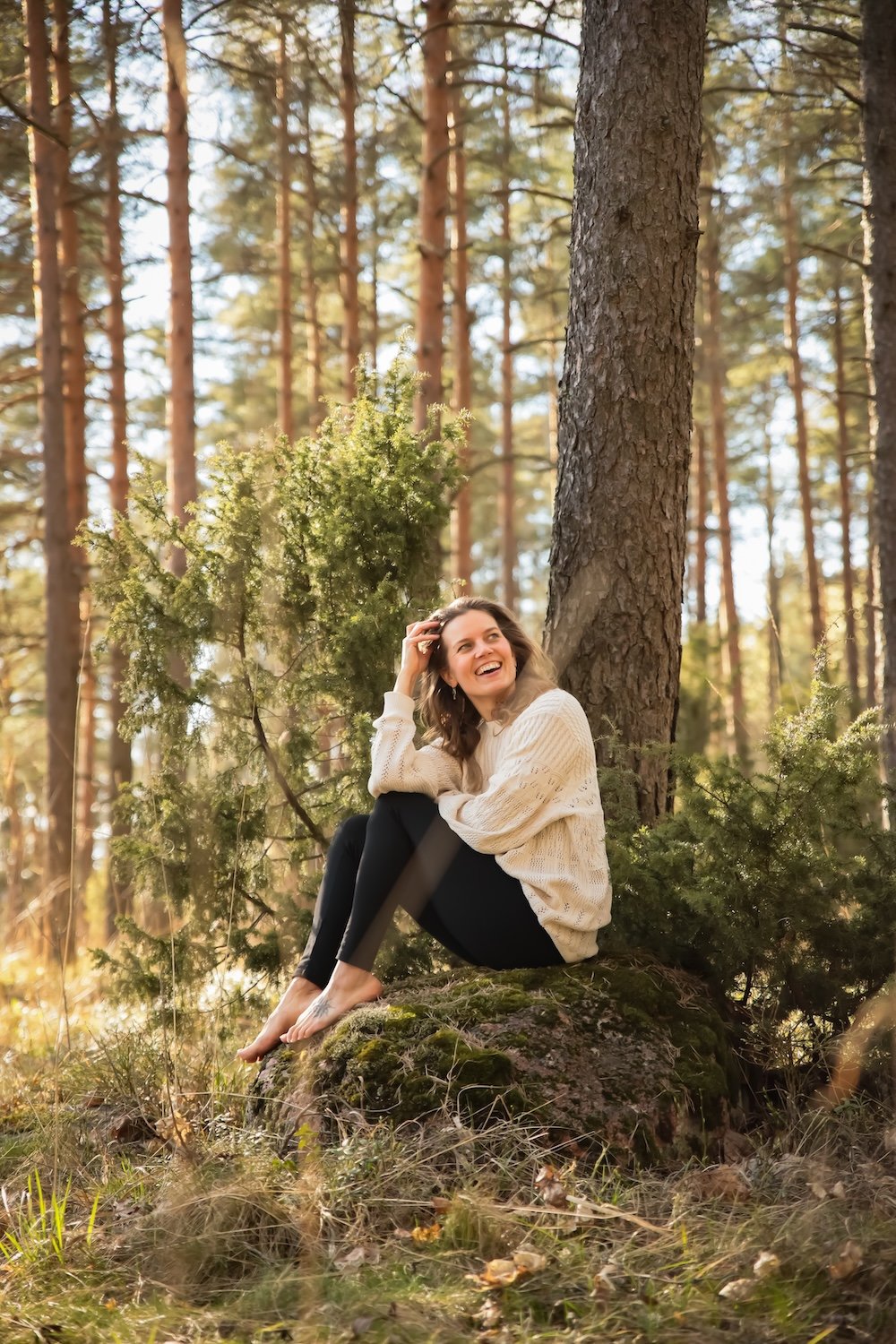 Äänimaljarentoutus ohjaaja Miila Rahomäki istuu soraisella kalliolla metsän keskellä, ympärillään vihreitä puita ja kasveja, taustalla puita ja sininen taivas.