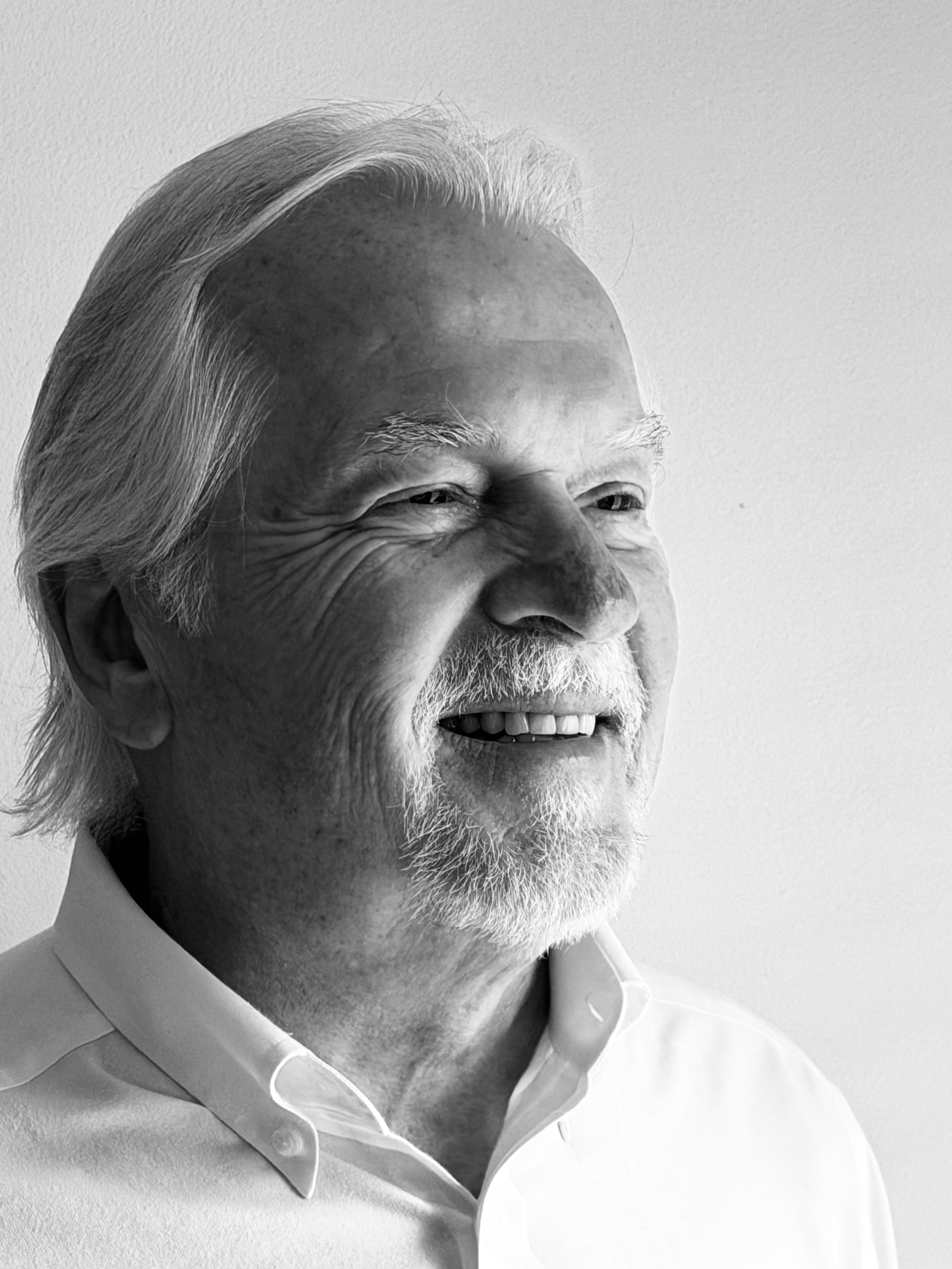 Close-up of a smiling elderly man with gray hair and beard, wearing a white shirt, against a plain background.