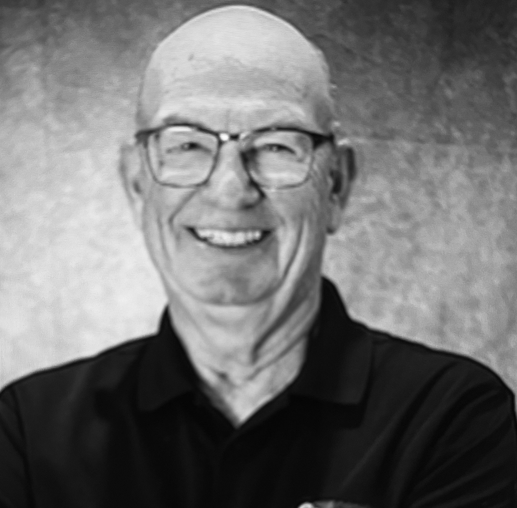Black and white photo of an elderly man smiling, wearing glasses and a cap, with a dark background.
