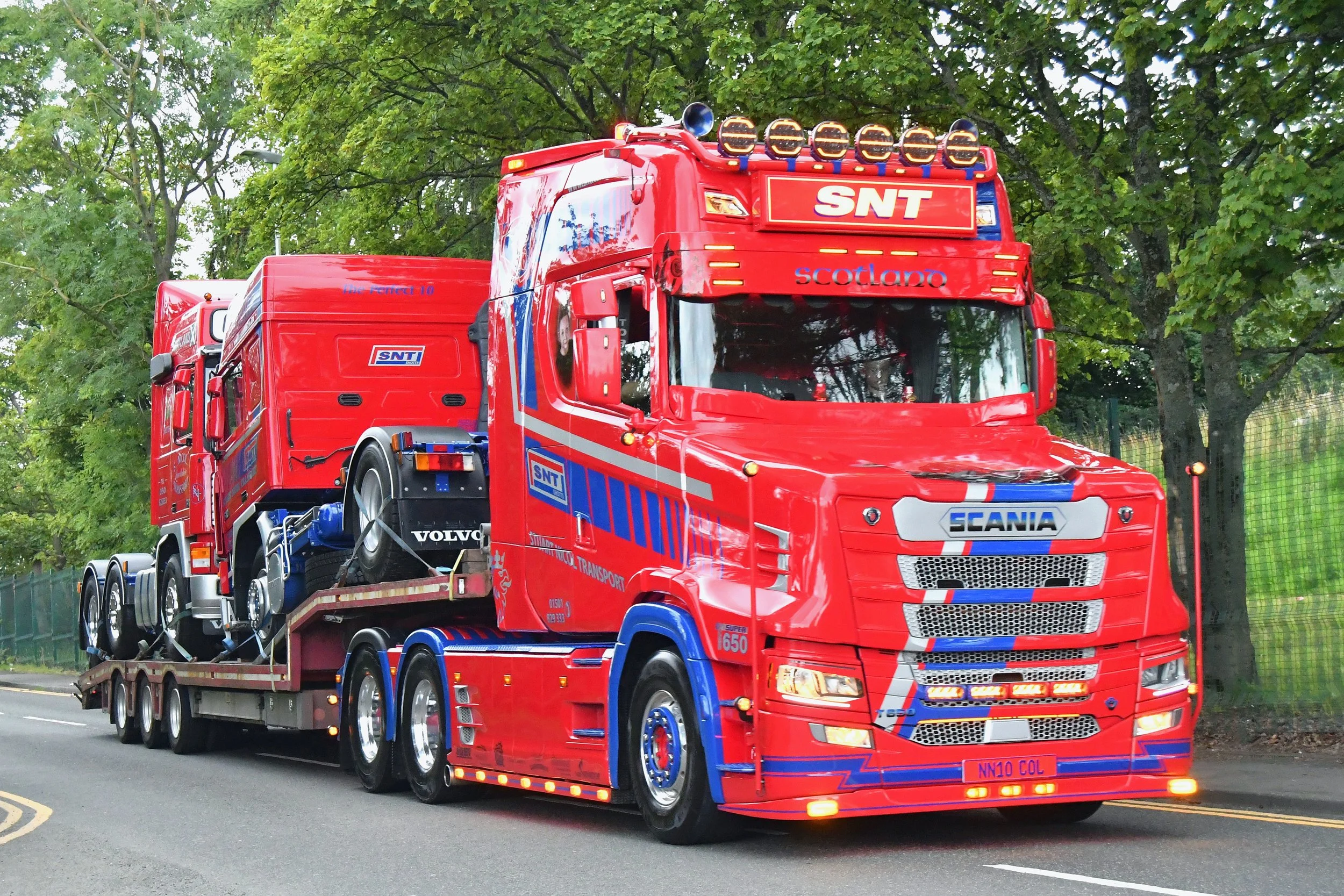 A red Scania truck with the words "SNT Scotlano" on the front, carrying a load of smaller red vehicles, on a road with green trees in the background.