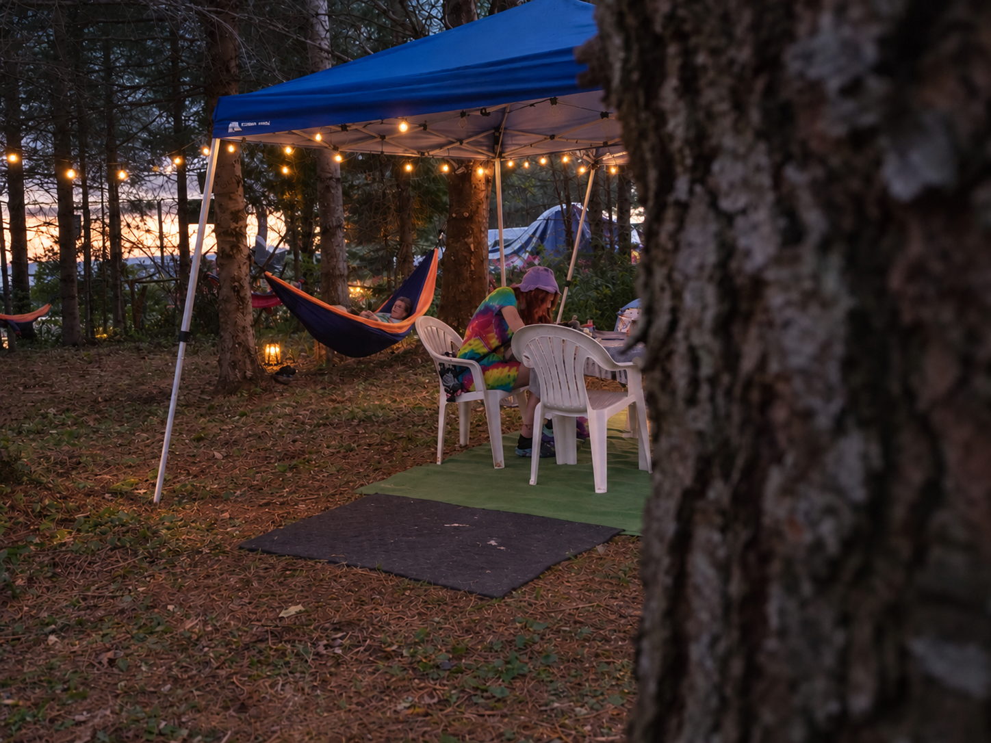 Camping scene at dusk with a canopy, string lights, a person sitting at a table, another person resting in a hammock, surrounded by trees and tents.