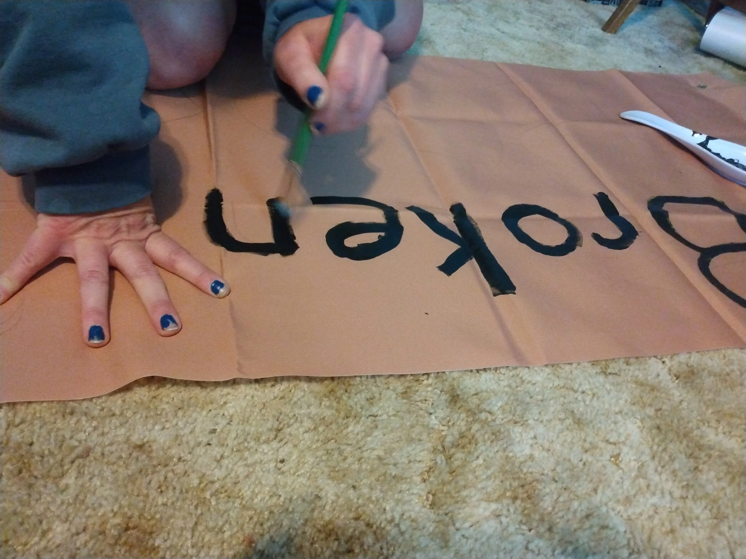 A brown fabric rectangle. A woman's hands are shown painting the word "broken' with black paint.