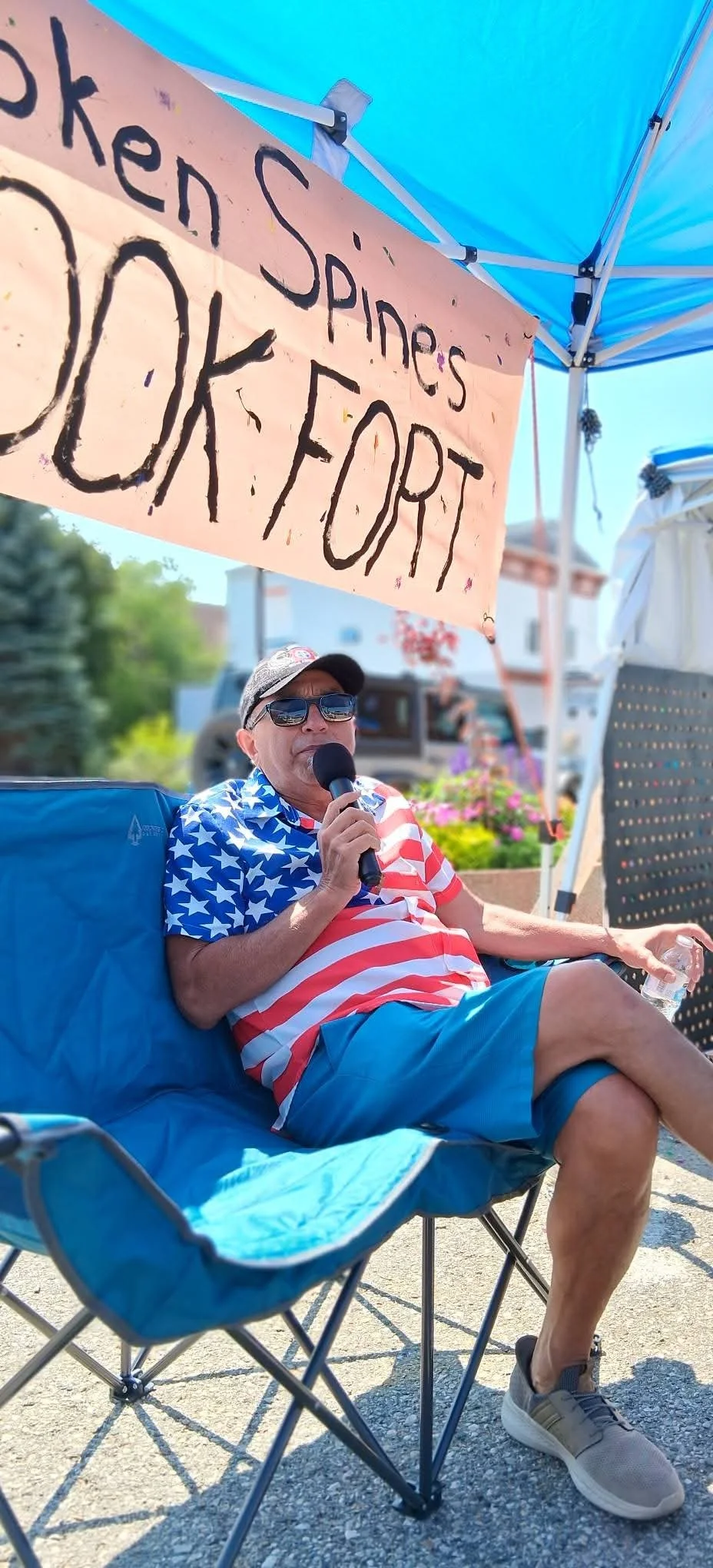 A man sitting in a folding chair under a blue canopy at an outdoor event, wearing sunglasses and patriotic clothing featuring the American flag pattern, holding a microphone, with a sign above him that reads 'Oregon Speaker's OK For It'.