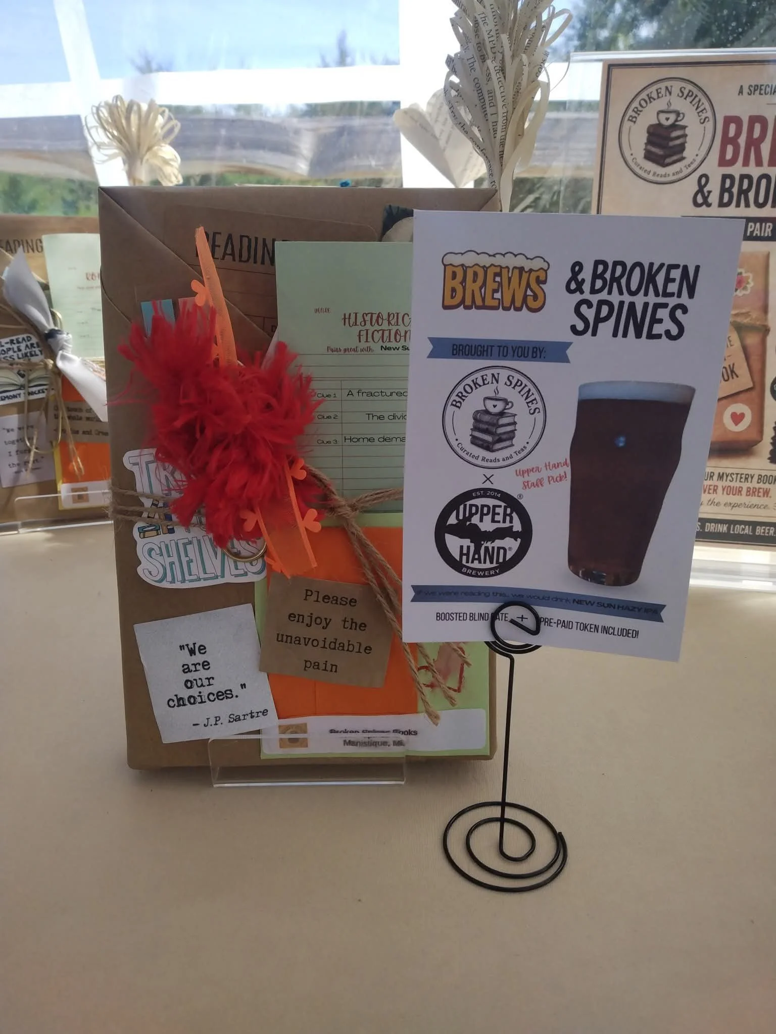 A display on a table with various items including a paper sign for 'Brews & Broken Spines', a paper bag with a red feather and orange ribbon, informational papers, and a small black spiral stand. The background shows a window with a blurry outdoor sc