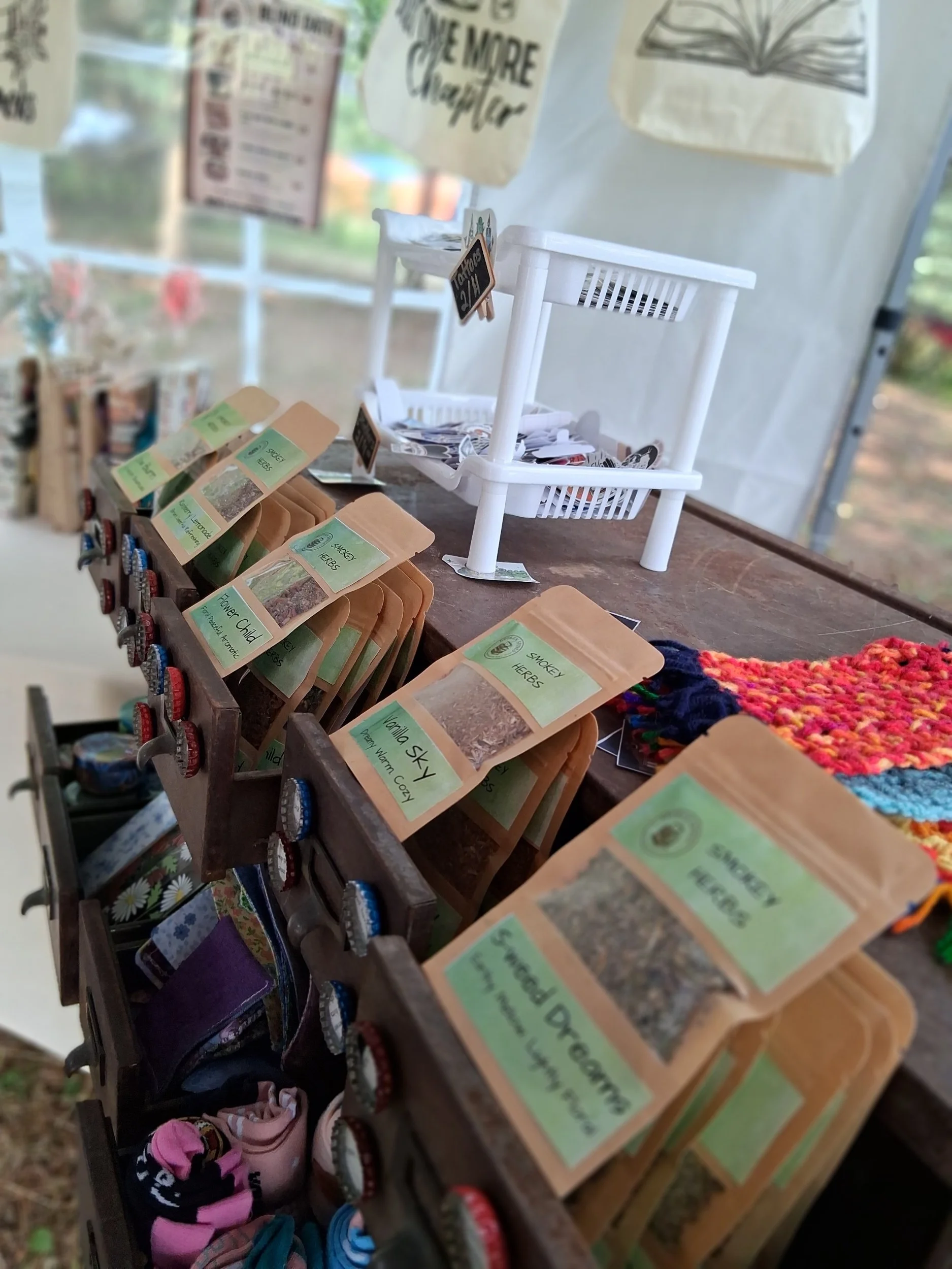 A display table with small paper bags labeled with different types of herbs, including 'Smokey Herbs', 'Vanilla Sky', 'Sweet Leaf', and 'Flower Child'. The table also features a small white multi-tiered stand with more herbs and related items, a colo