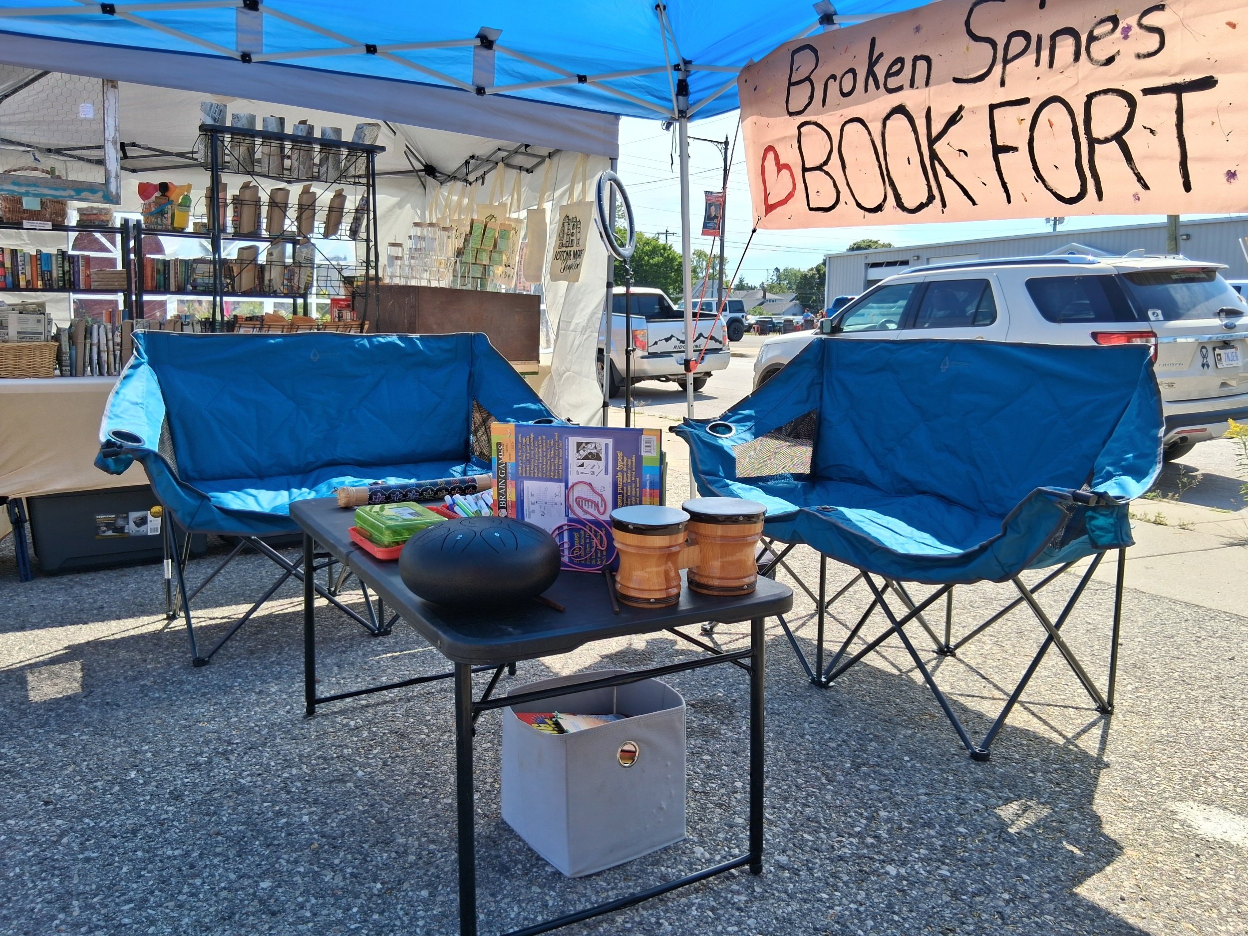 A booth at an outdoor market with a sign that says 'Broken Spines Book Fort'. The booth has books, containers, and various paper items on display. There are chairs with blue cushions in front of the booth, and cars parked in the background on a sunny day.