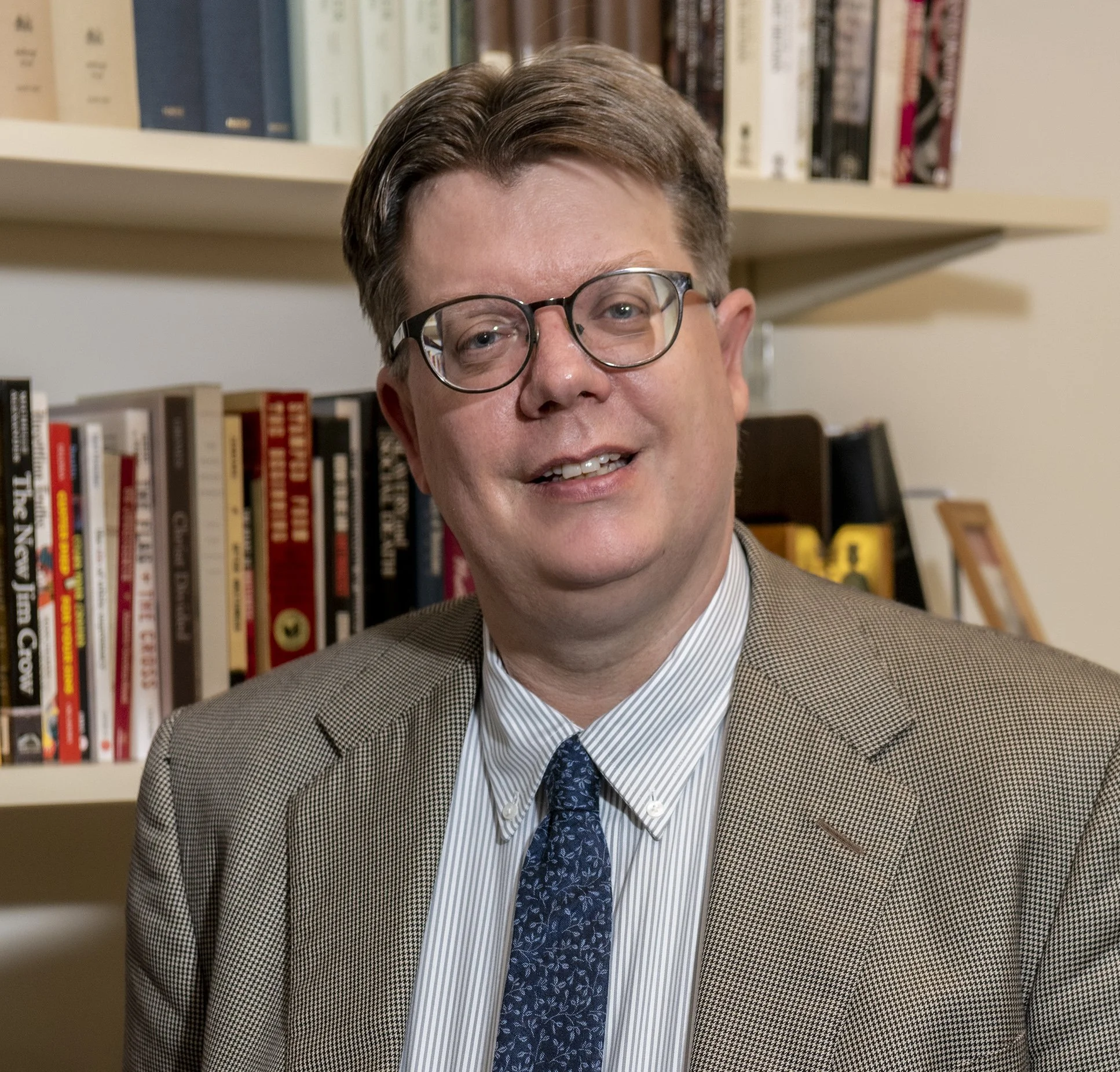 A man with glasses and brown hair wearing a tan houndstooth blazer, light blue dress shirt, and dark patterned tie, standing in front of a bookshelf filled with books.