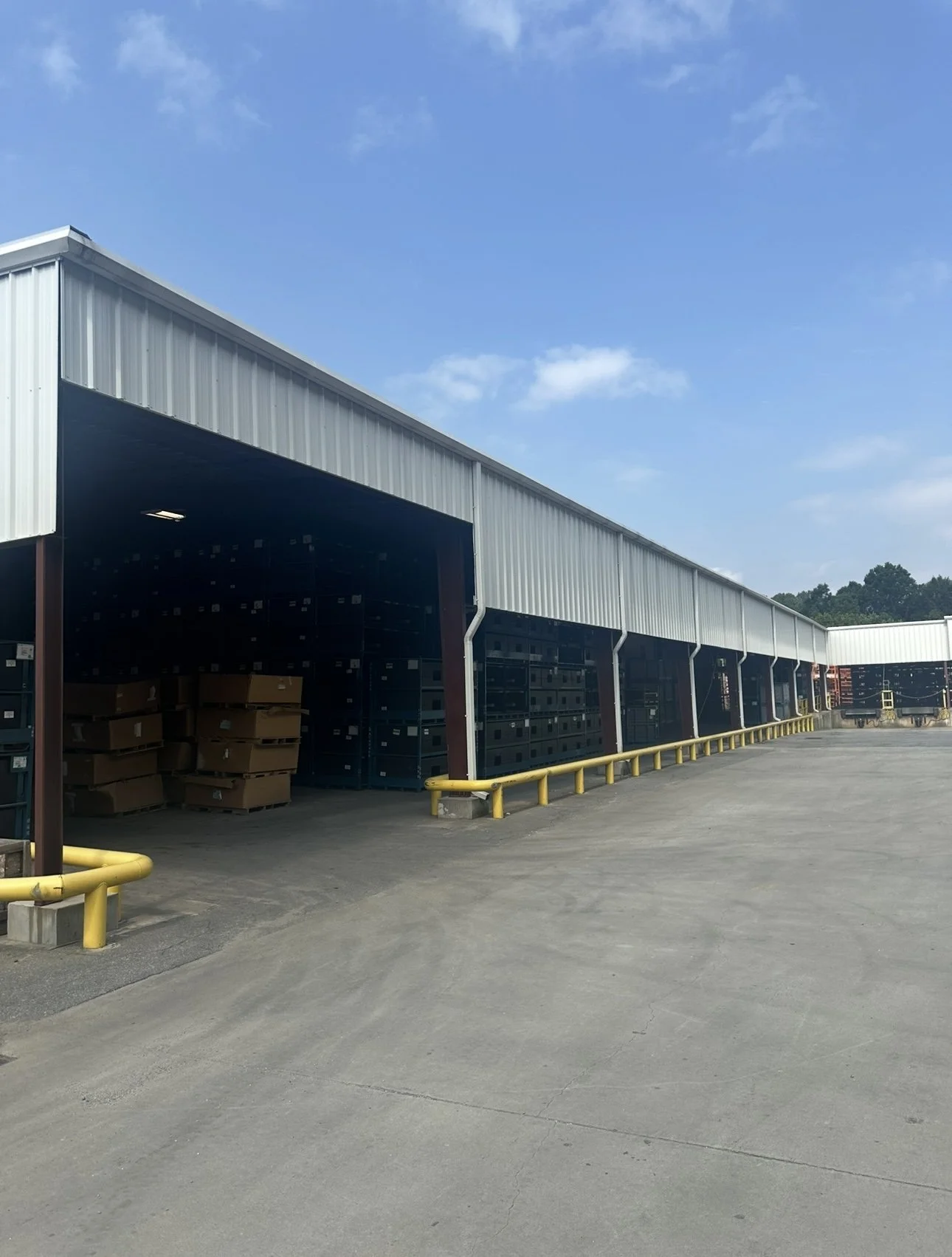 A warehouse or storage facility with open bays, containing boxes and pallets, with a clear blue sky overhead.
