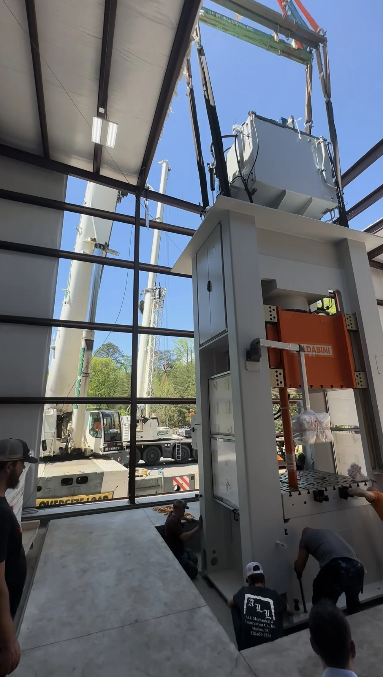 Workers installing heavy machinery inside a building under construction, with construction equipment visible outside through large open walls.