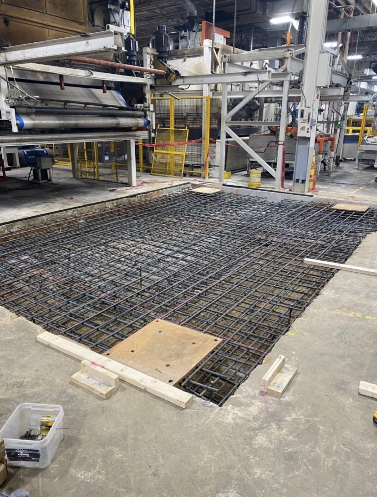 Empty steel rebar grid laid on the concrete floor, preparing for concrete pouring at a construction site, with construction machinery and safety barriers in the background.