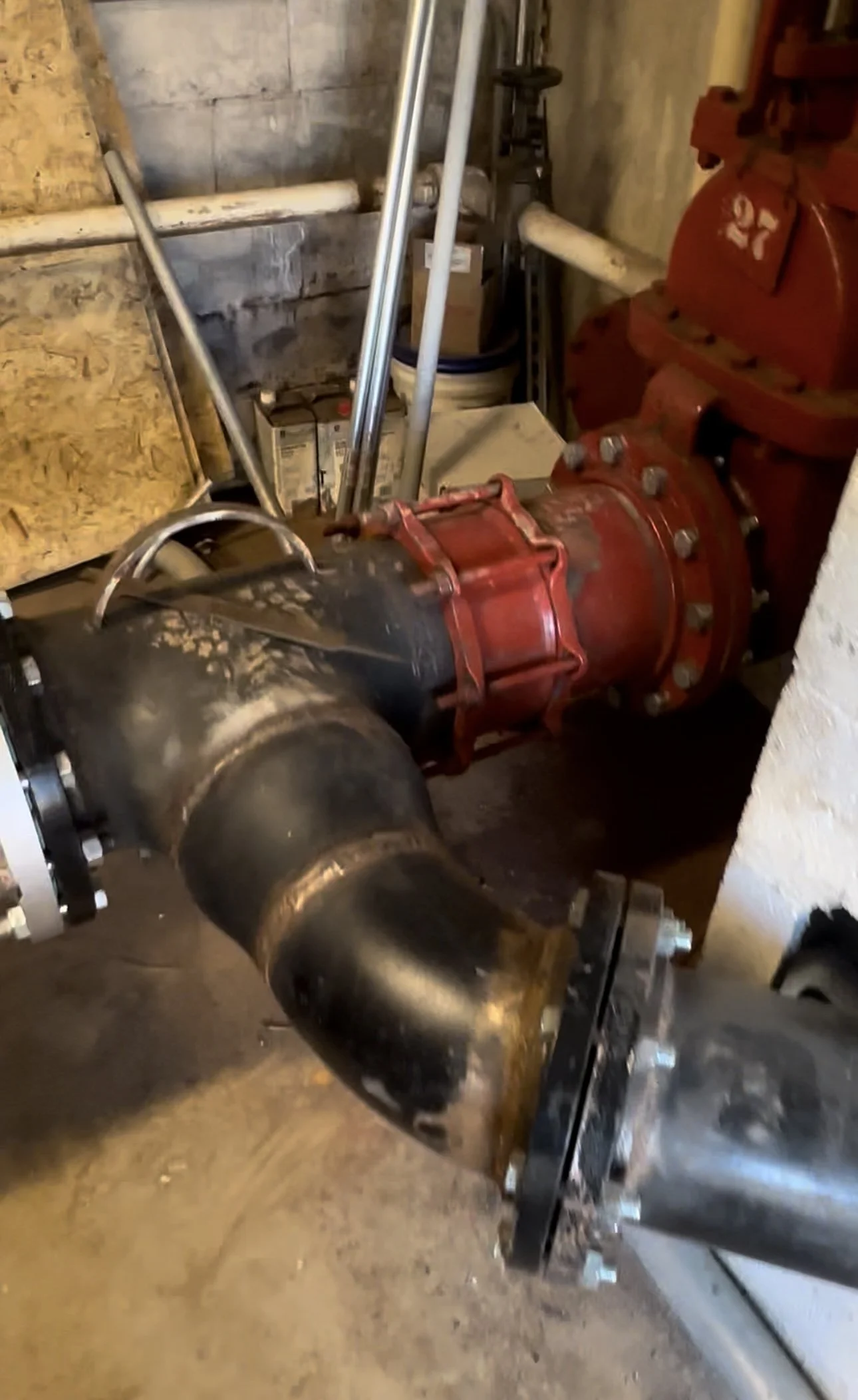 A large industrial pipe system with a red valve and metallic piping, set against a concrete wall with miscellaneous tools and objects nearby.