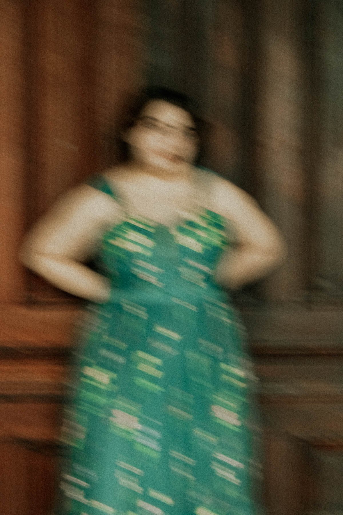 A woman standing with hands on hips, wearing a blue dress with green and white patterns, in front of a dark curtain. The image is blurry.