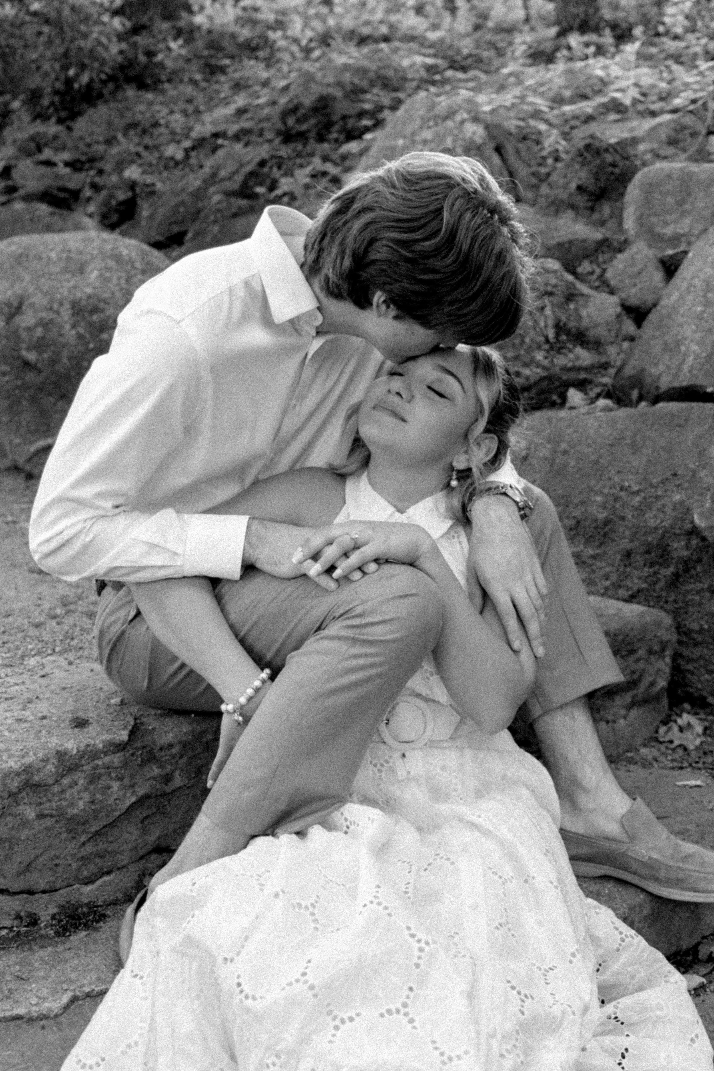 A young man and woman sit on rocks outdoors; the man leans down to kiss the woman's forehead as she smiles with her eyes closed. The woman wears a dress with eyelet details, and the man wears a button-up shirt.