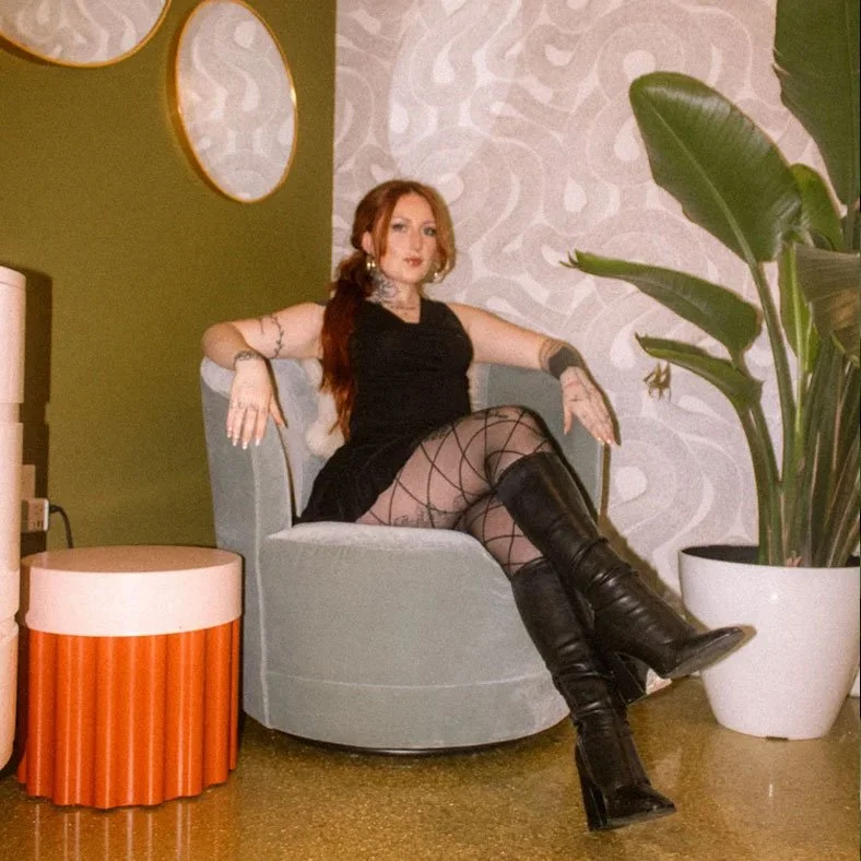 A woman sitting on a gray armchair in a room with green and patterned white walls, large green plant in white pot nearby, orange cylindrical side table, dressed in black with fishnet stockings and black knee-high boots.