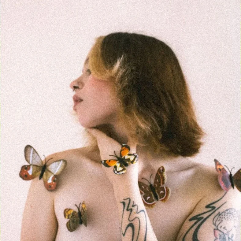 A woman with short, two-tone hair featuring blonde and brown shades, looking to the side. She has colorful butterfly tattoos on her shoulder and hand, and butterflies are perched on her shoulder, chest, and neck.