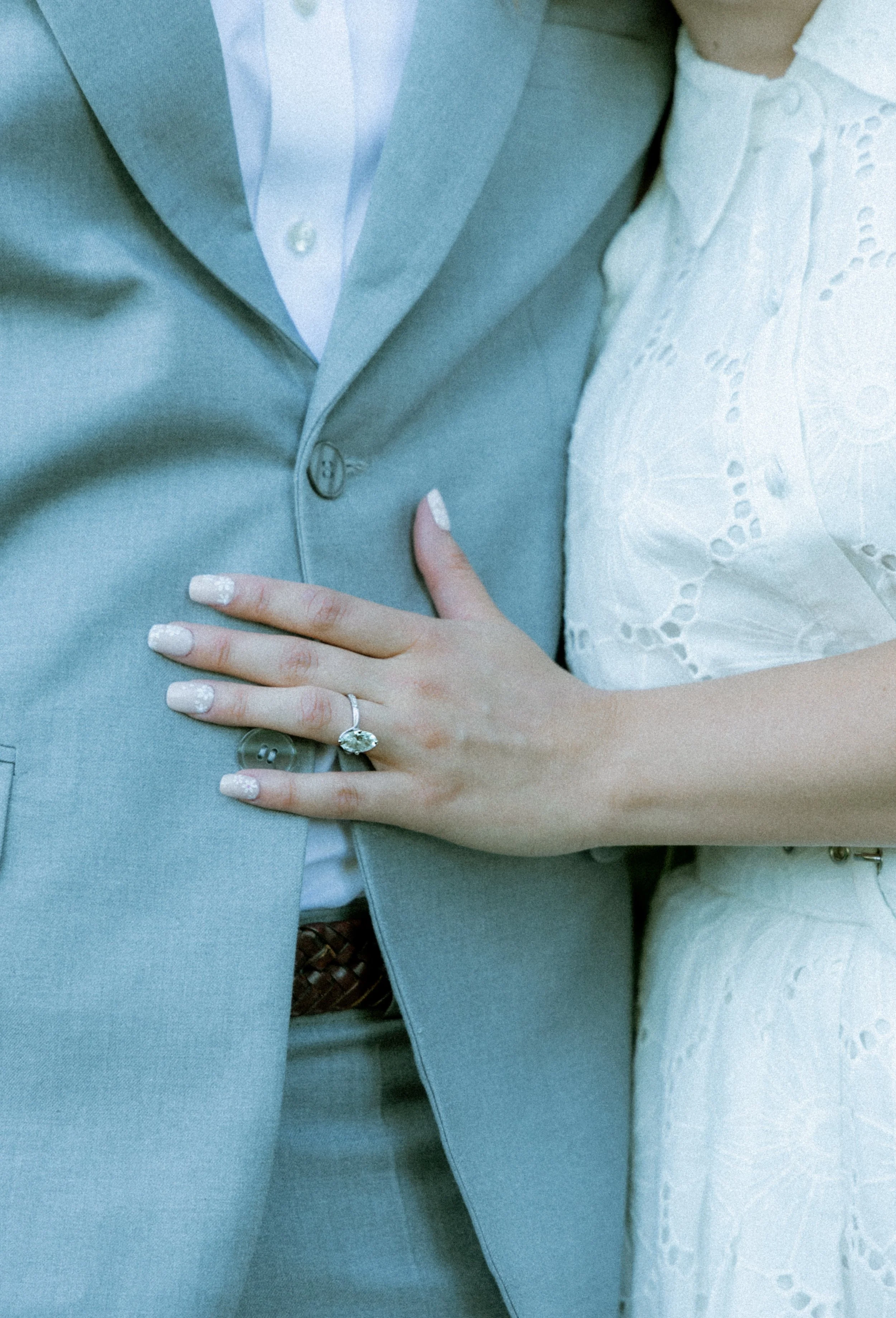 Close-up of a bride and groom, focusing on their torsos. The groom is wearing a light blue suit with a white shirt, and the bride is in a white dress with eyelet embroidery. The bride's hand, showing a wedding ring, is resting on the groom's chest.