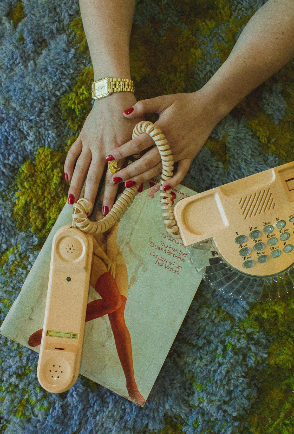 Close-up of a vintage peach-colored rotary telephone on top of a magazine, with a person's hands holding it. The person has red nails, wears a gold watch, and a cream-colored spiral bracelet. The background is a textured, multicolored rug.