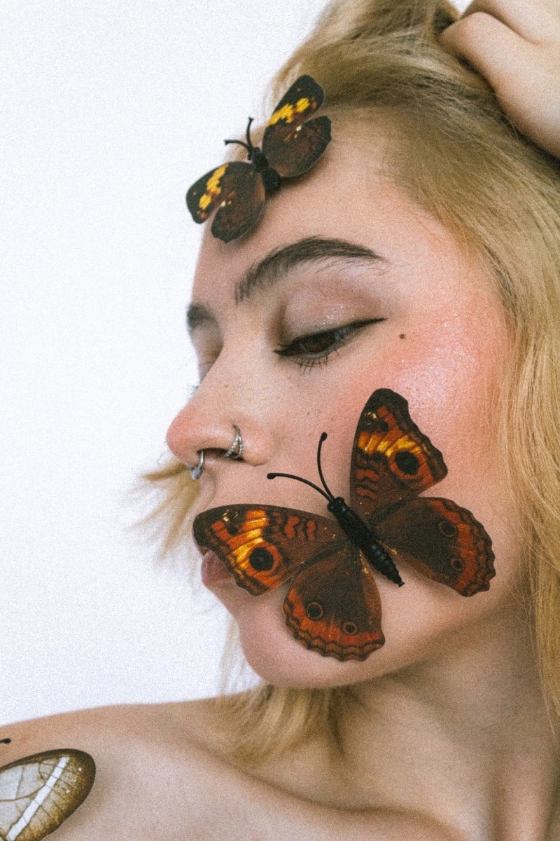 A woman with blonde hair and a septum piercing has three large orange and brown butterflies on her face and hair, against a plain white background.